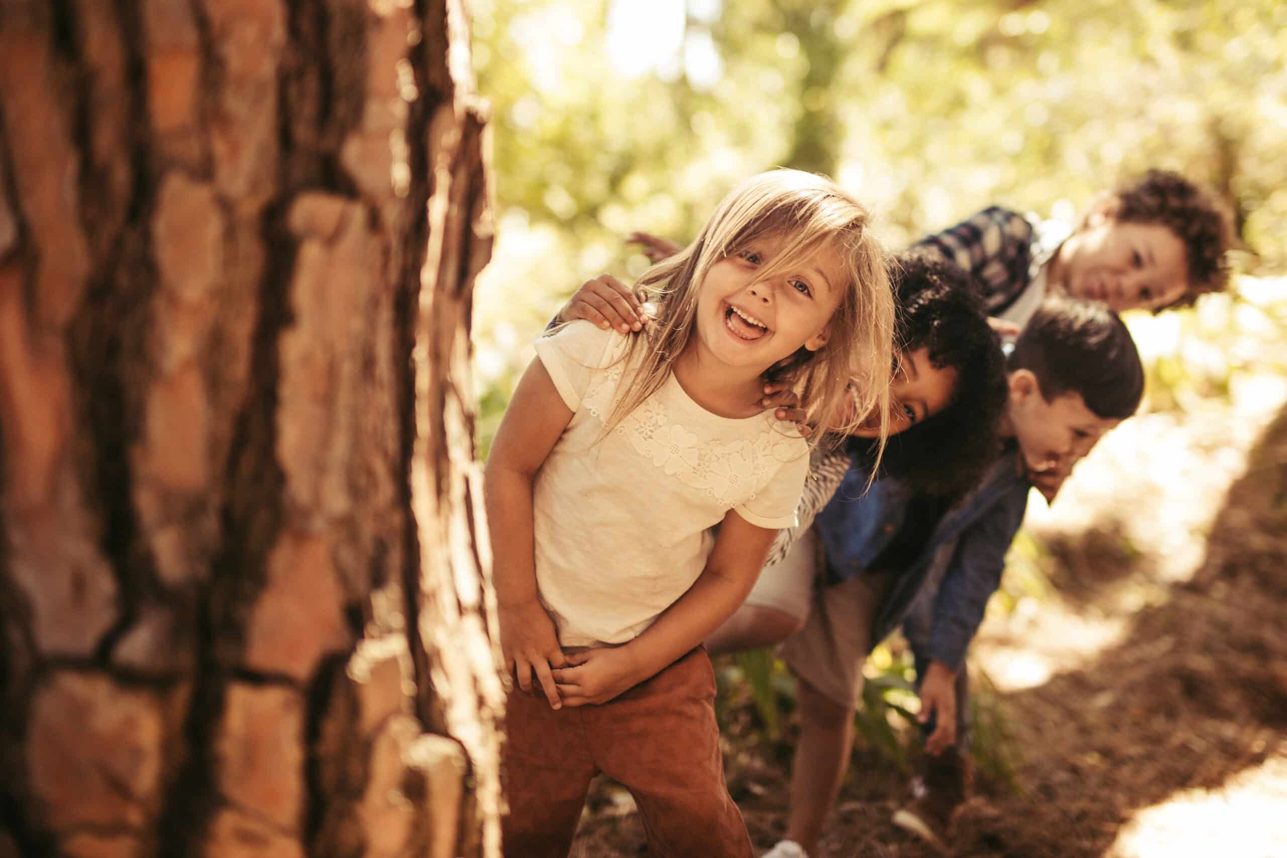 Group of kids standing in row and peeking behind a tree outdoors. Children playing hide and seek in a park.