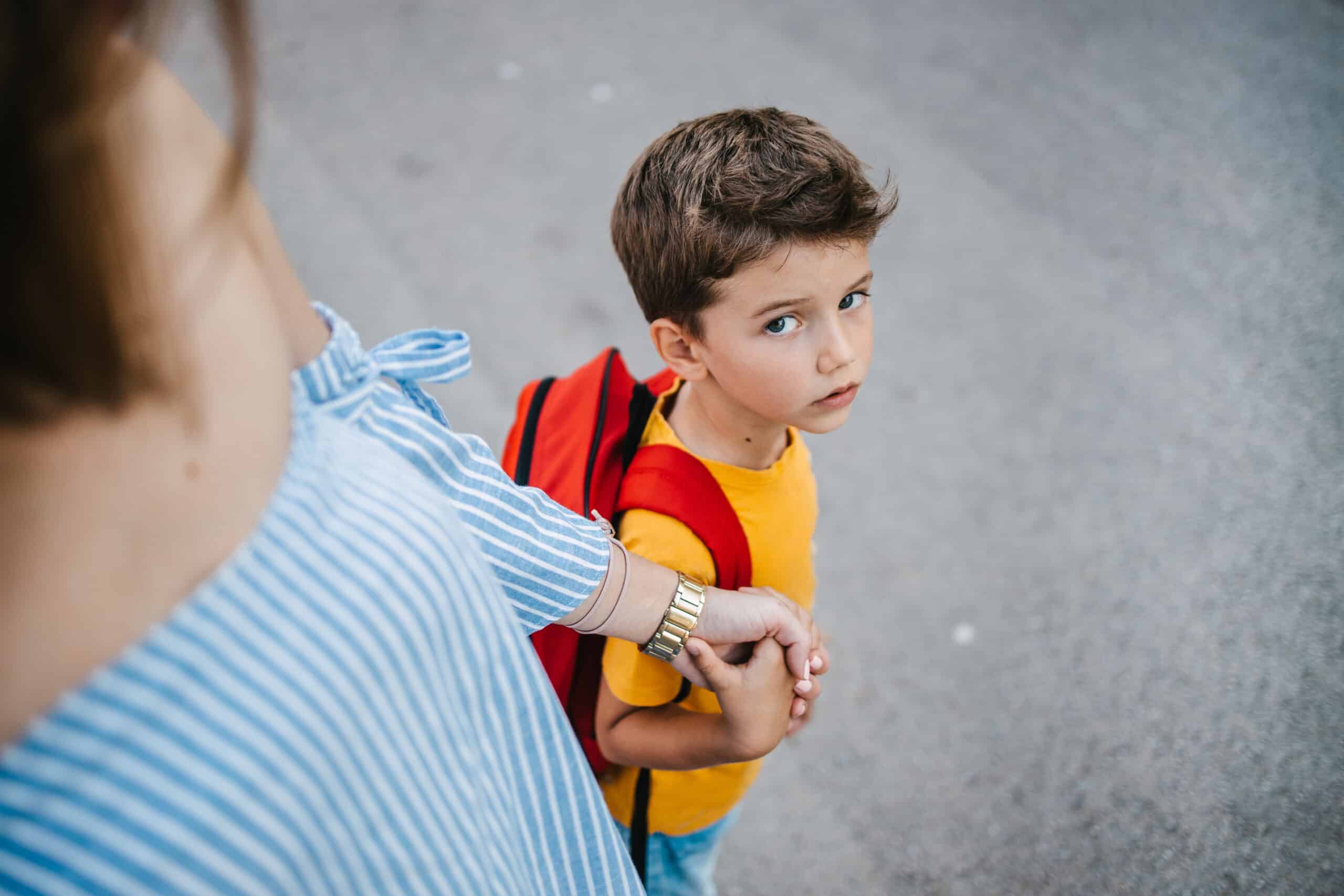 A son with a backpack and his mother holding his hand sending him to school on the first day of school and the schoolboy is looking at the camera