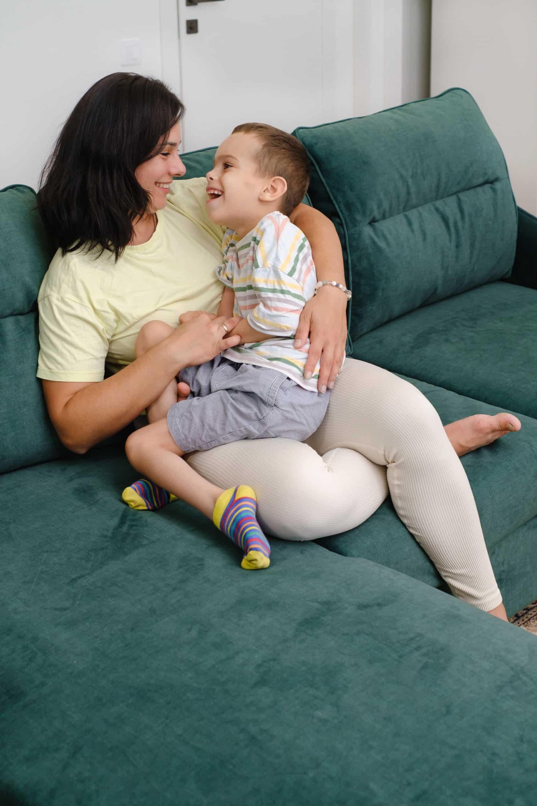 Mother holding child, living with disability on sofa Woman hugging boy with cerebral palsy at home Mother having fun, taking care of boy that has chronical health condition Care and love at home