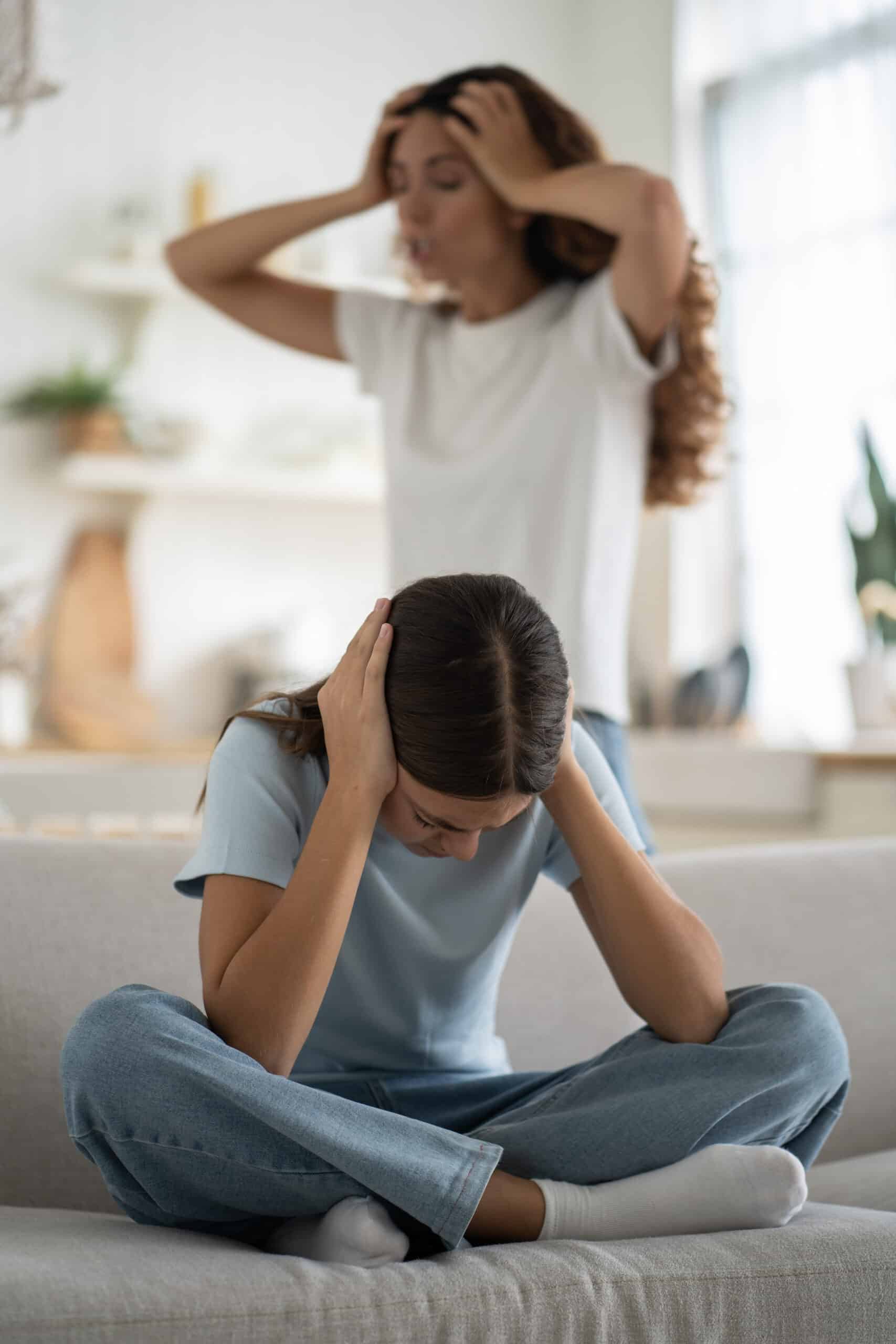 Depressed teenage girl covering ears with hands while her frustrated angry mother yelling on background. Toxic parent scolding daughter at home. Adolescent behavior. Psychological abuse of child