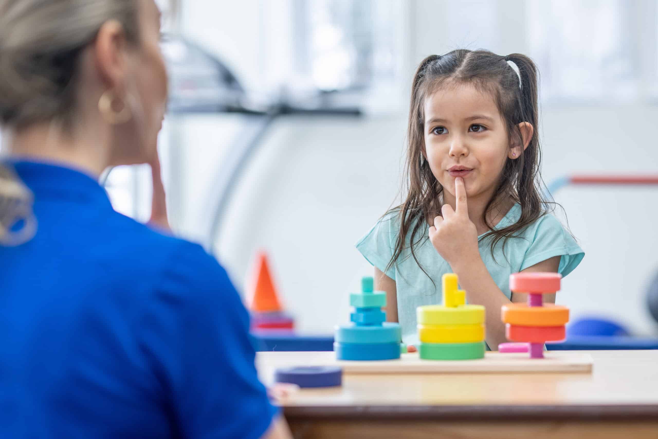 A female therapist sits with a sweet little girl as a table as they work on her speech together.  The Therapist is dressed professionally and is pointing to her mouth to help show the little girl how to form her words.