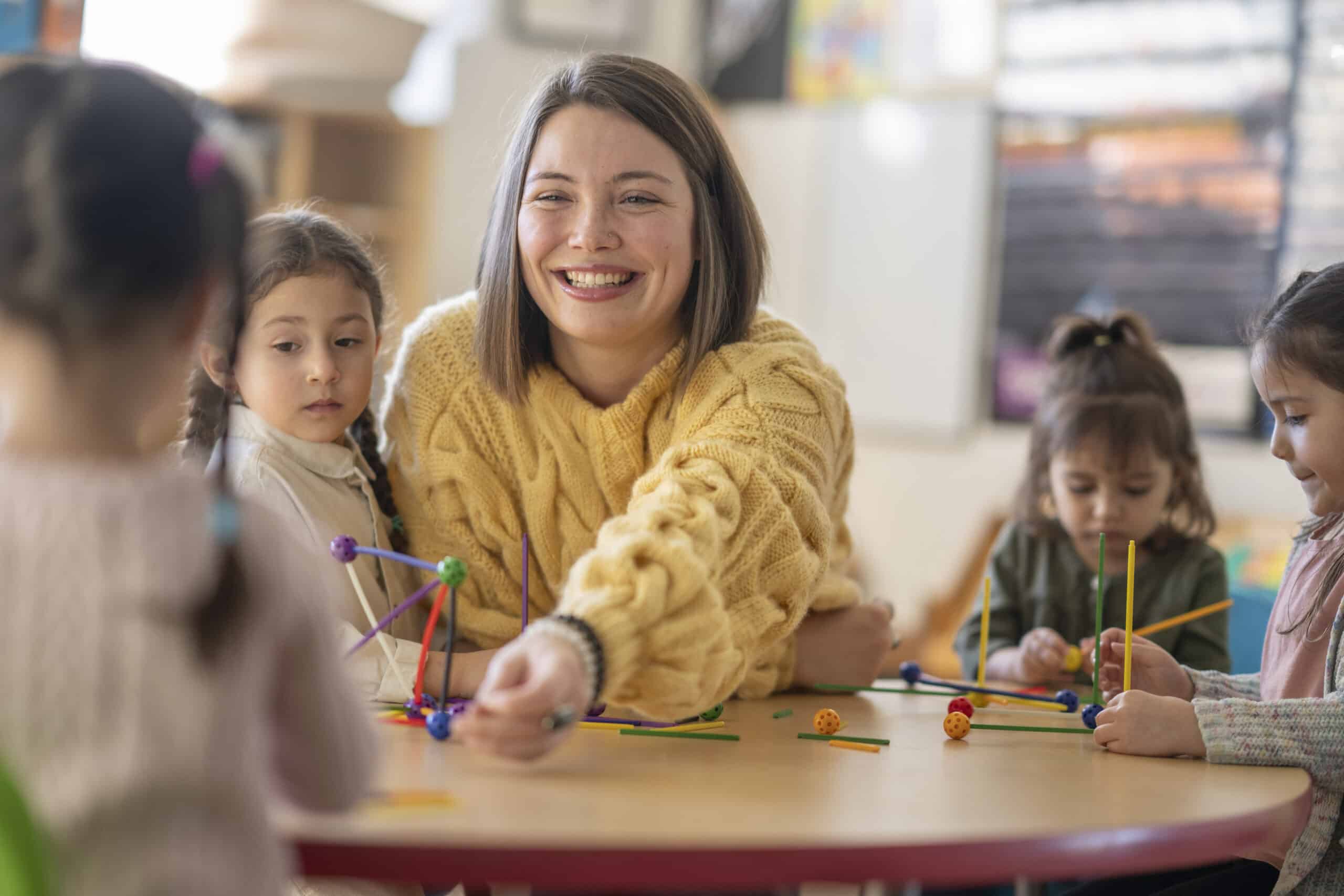 A small group of Kindergarten students sit with their female teacher as they work away building structures.  They are each dressed casually as they work on their projects and talk amongst themselves.