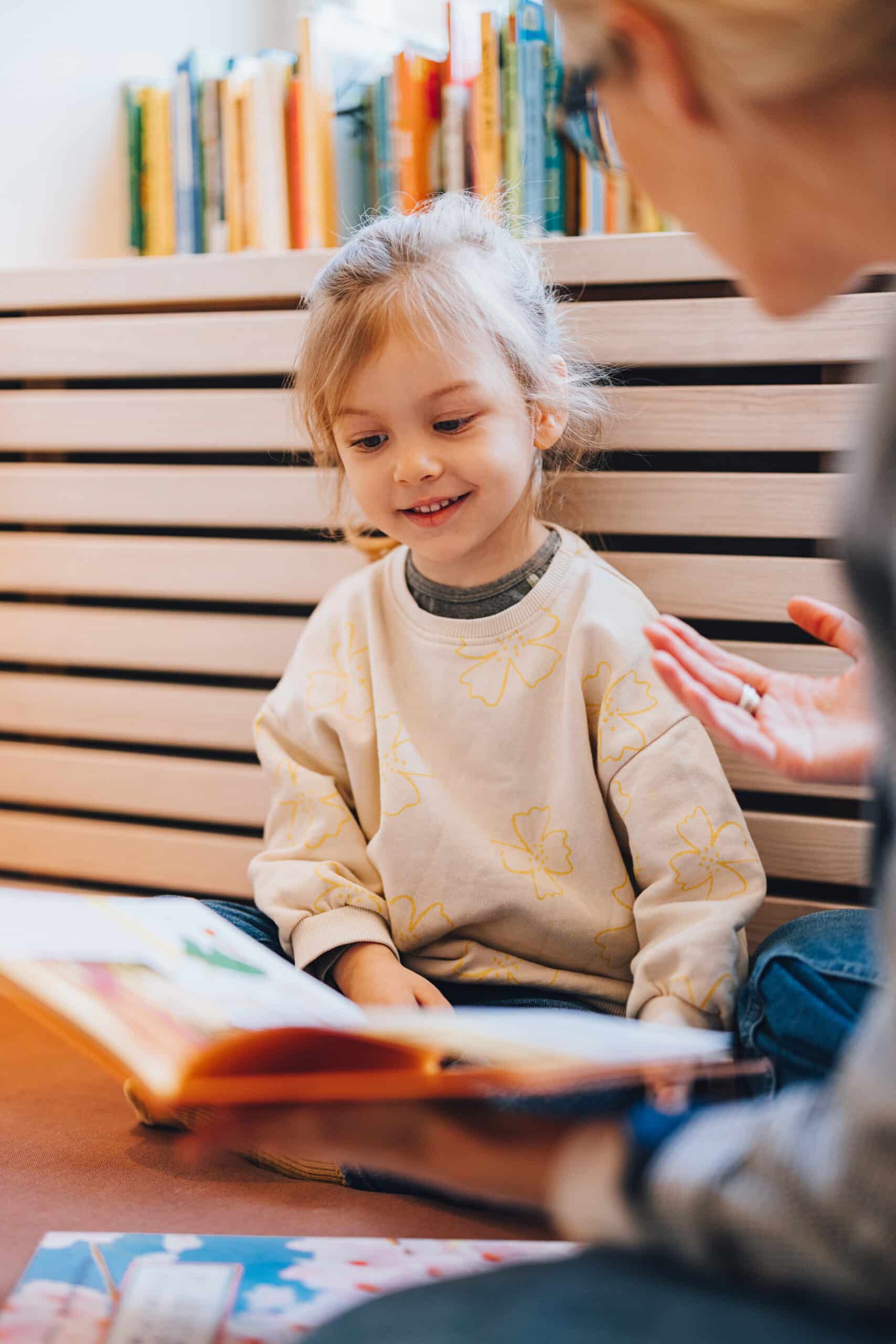 Close up shot of a cute young girl sitting on the floor in the library. Unrecognizable woman is holding a book and showing or explaining something to her. She is looking down at the book.